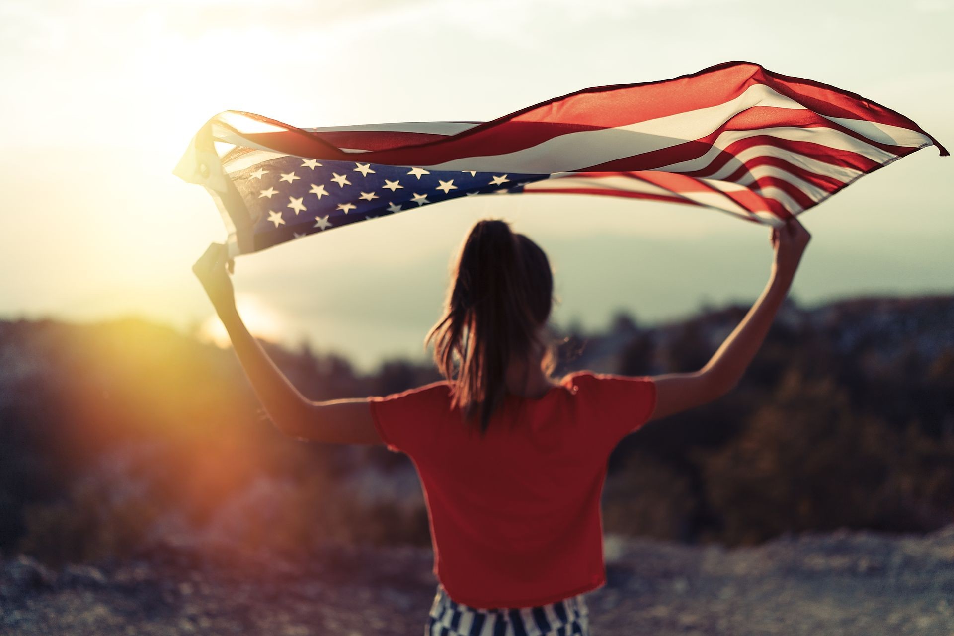Child girl is waving American flag on top of mountain at sky background. Sunset time. USA resident, US citizen. Immigration concept